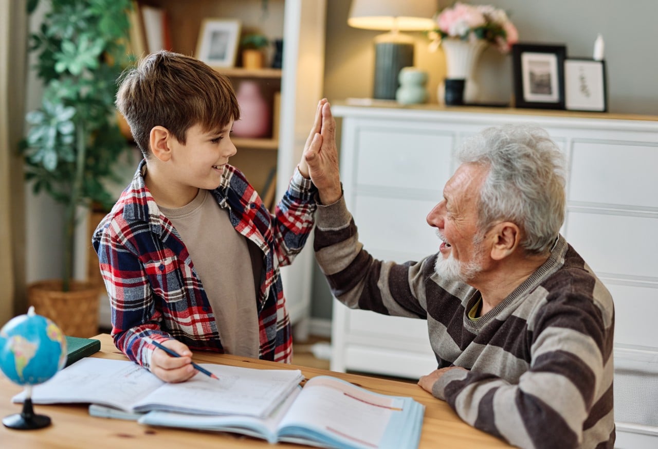 A Nurturing Alternative to Mainstream Education Smiling elderly man and young boy high-five while doing homework at a table.