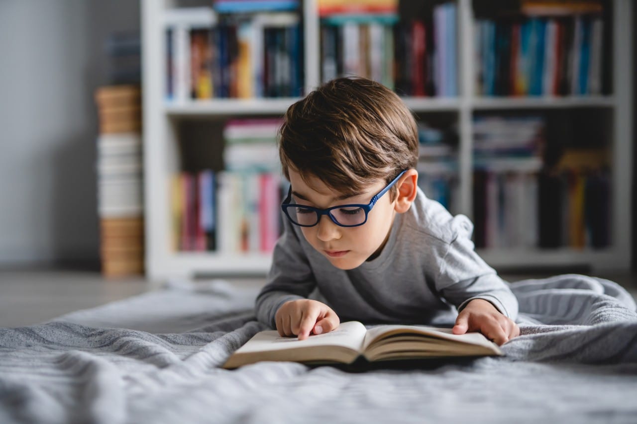 A young boy wearing glasses reads a book while lying on the floor in front of a bookshelf.