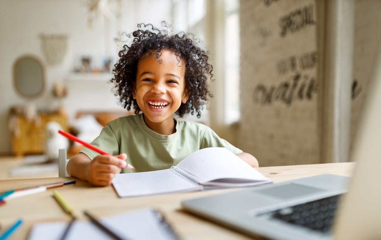 Smiling child sitting at a desk, holding a pencil and writing in a notebook.