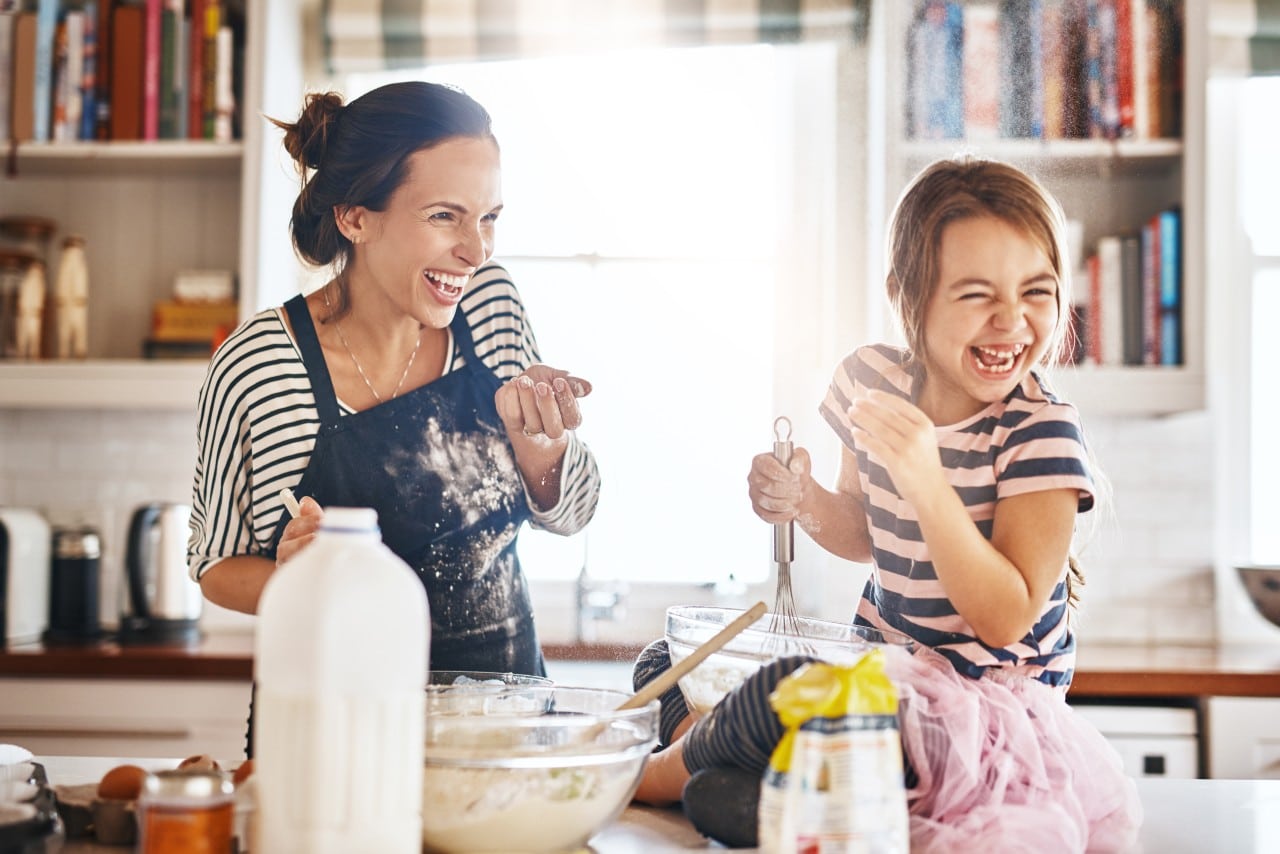 Education That Feels Relevant to Real Life A woman and a girl laugh together while baking in a bright kitchen.