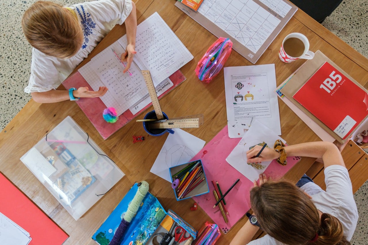 Two children doing schoolwork at a table with notebooks, rulers, colored pencils, and folders.