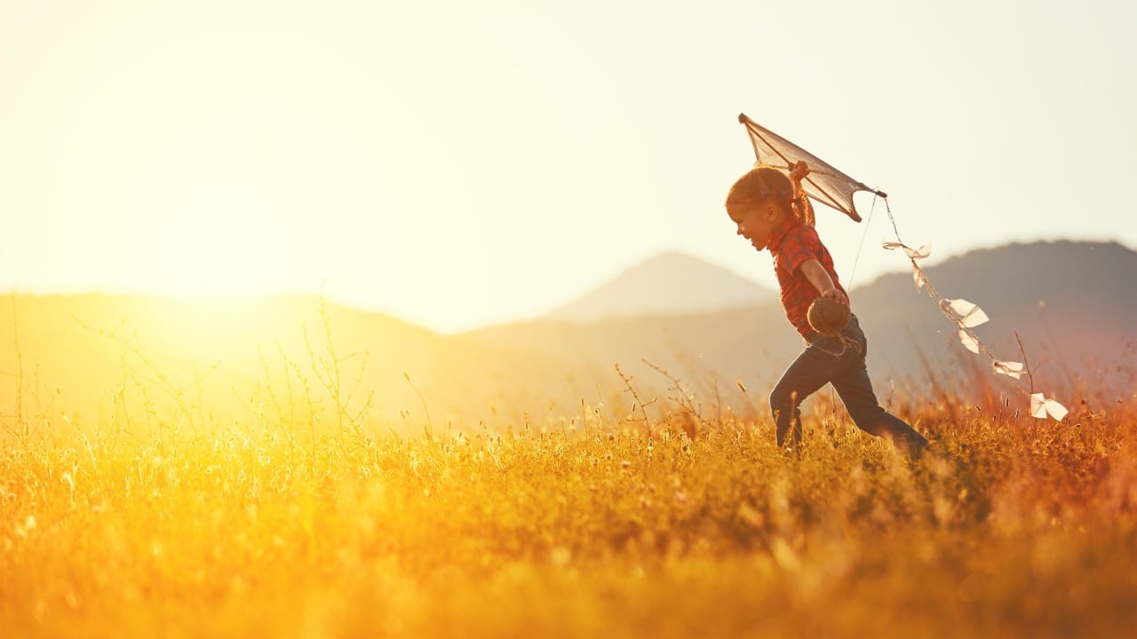 Child running with a kite in a sunny field with mountains in the background.