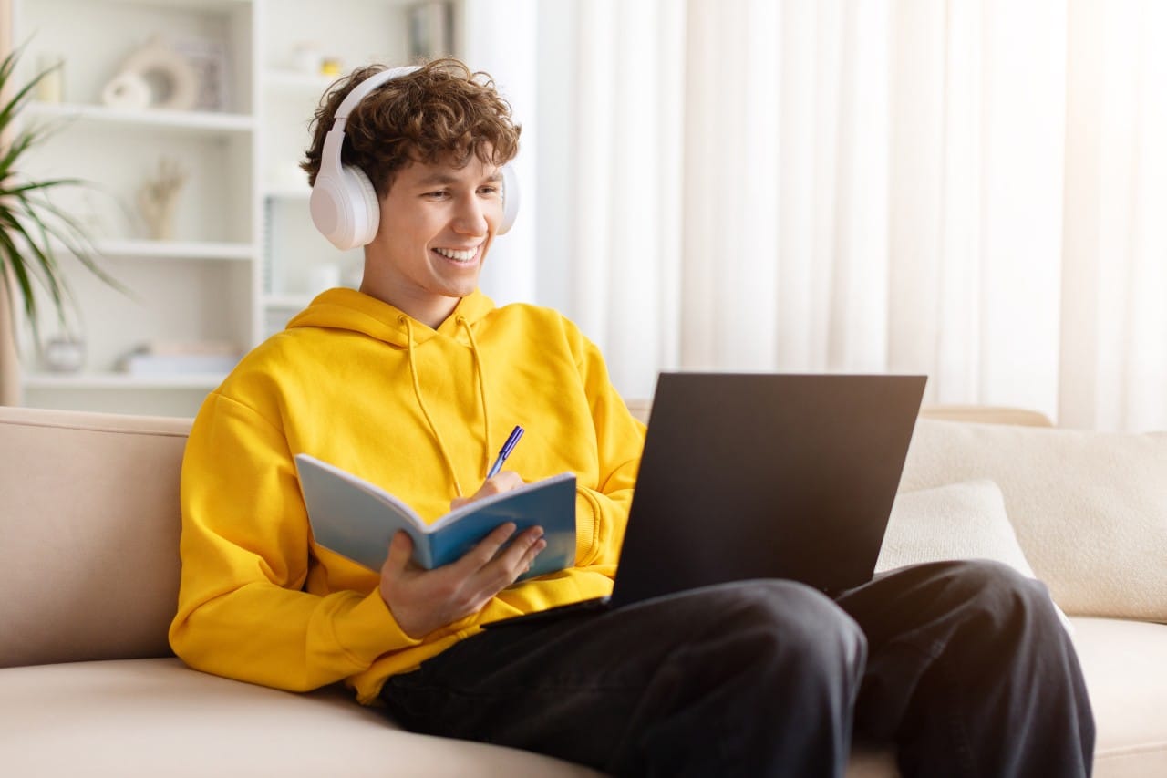 Smiling person with headphones studying on a laptop and writing in a notebook on a couch.